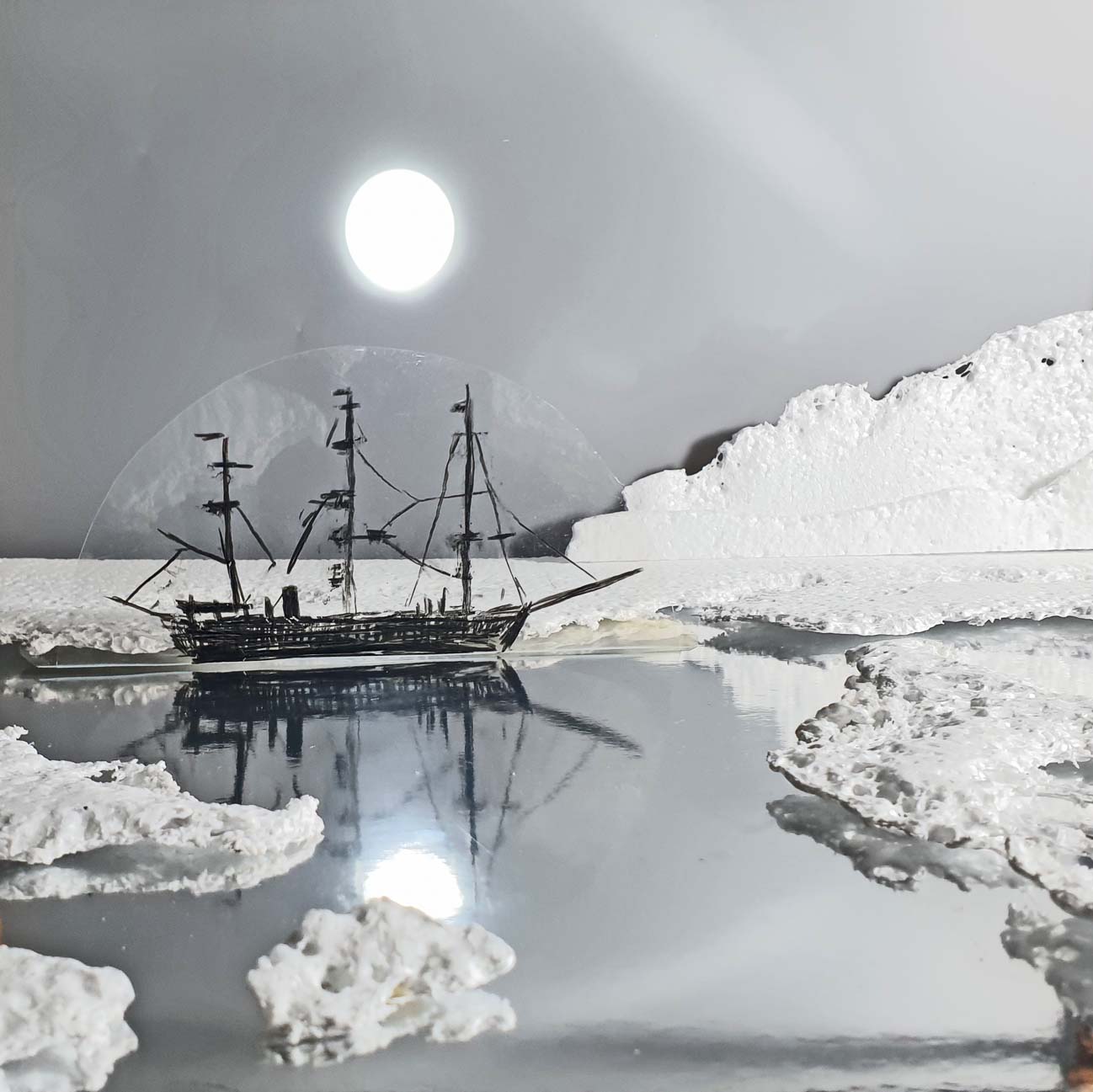 Glowing white moon imgae appears above a drawing of an old fashioned whaling ship reflected in a mirrored surface with icebergs and snow made of packaging